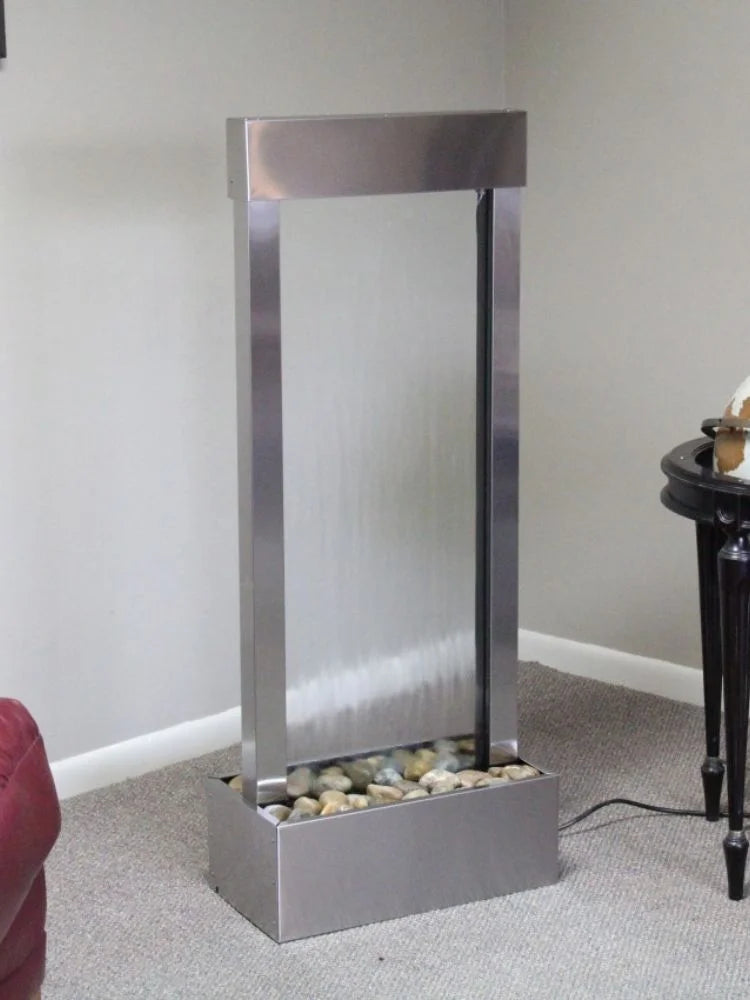 A modern rectangular waterfall floor lamp with a brushed stainless steel frame, a frosted glass waterfall panel, and a base filled with decorative river stones, shown in a home setting on a carpeted floor.