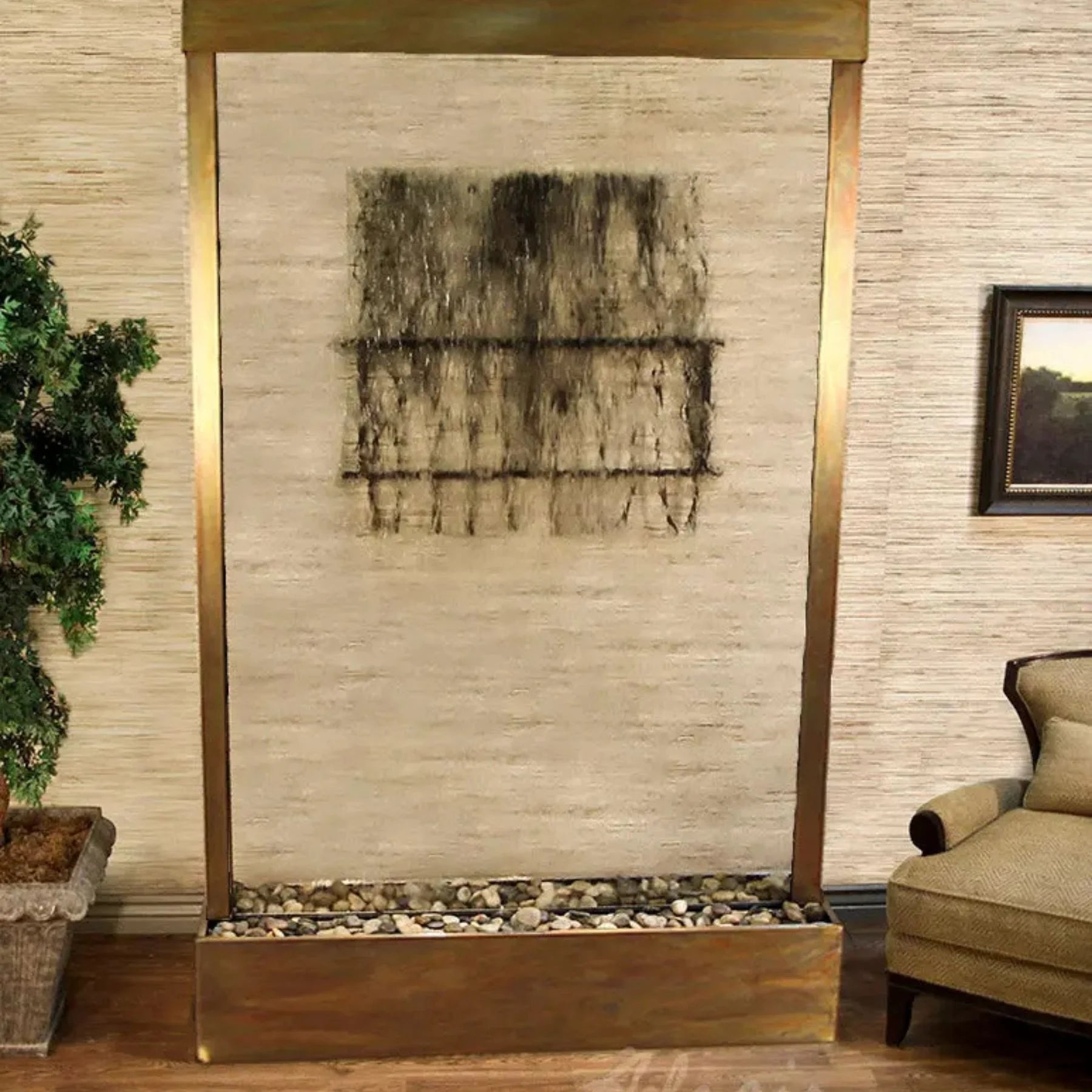 A large rectangular wall-mounted art piece with a gold-toned frame, featuring a textured abstract black and gray design on a beige background, displayed in an indoor room setting with a plant and armchair visible.