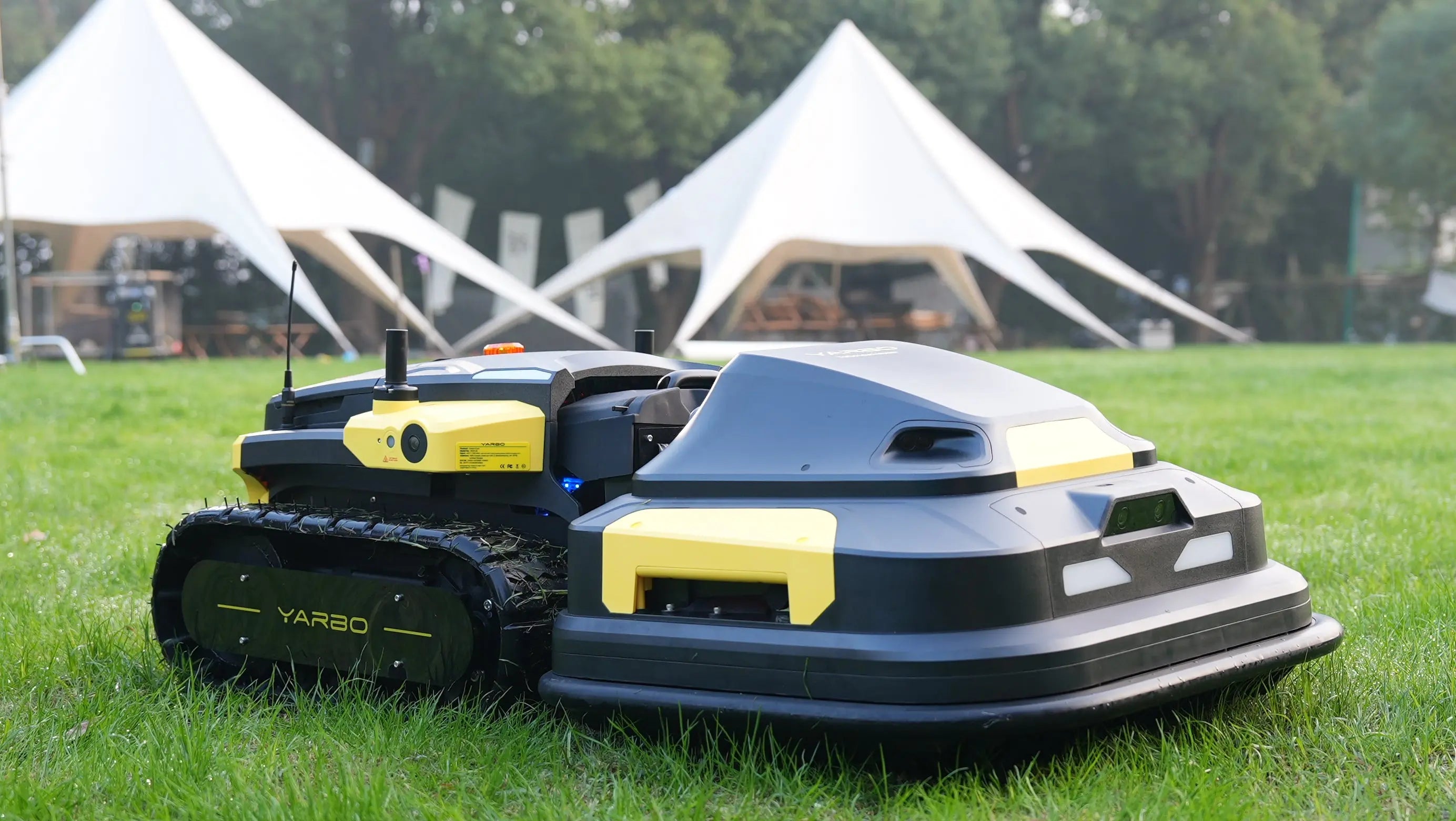 Robotic lawn mower on grass with tents in the background