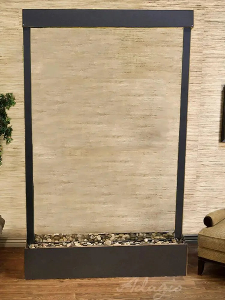 A rectangular freestanding wall fountain or decorative waterfall feature with a beige textured stone or plaster panel, dark charcoal metal frame, and a base filled with small decorative stones, shown in an indoor room setting.