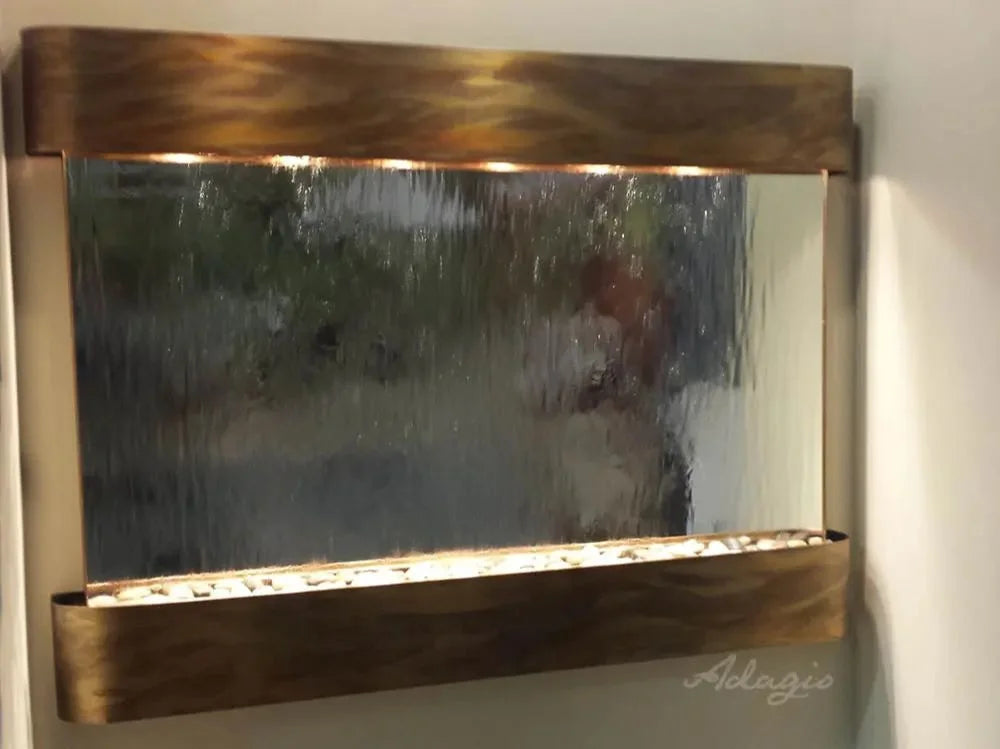 A rectangular wall-mounted mirror with a rustic bronze metal frame and integrated warm lighting, shown mounted on a white wall. The mirror surface reflects a blurred outdoor scene with greenery and a person, likely from a photo behind the camera.