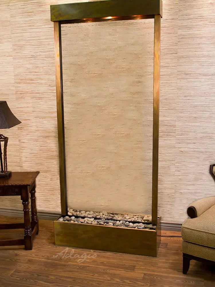 A freestanding rectangular wall-mounted waterfall feature with a textured beige stone or plaster face, framed in dark bronze metal, set in a furnished room with wood flooring, a side table, and part of an armchair visible.
