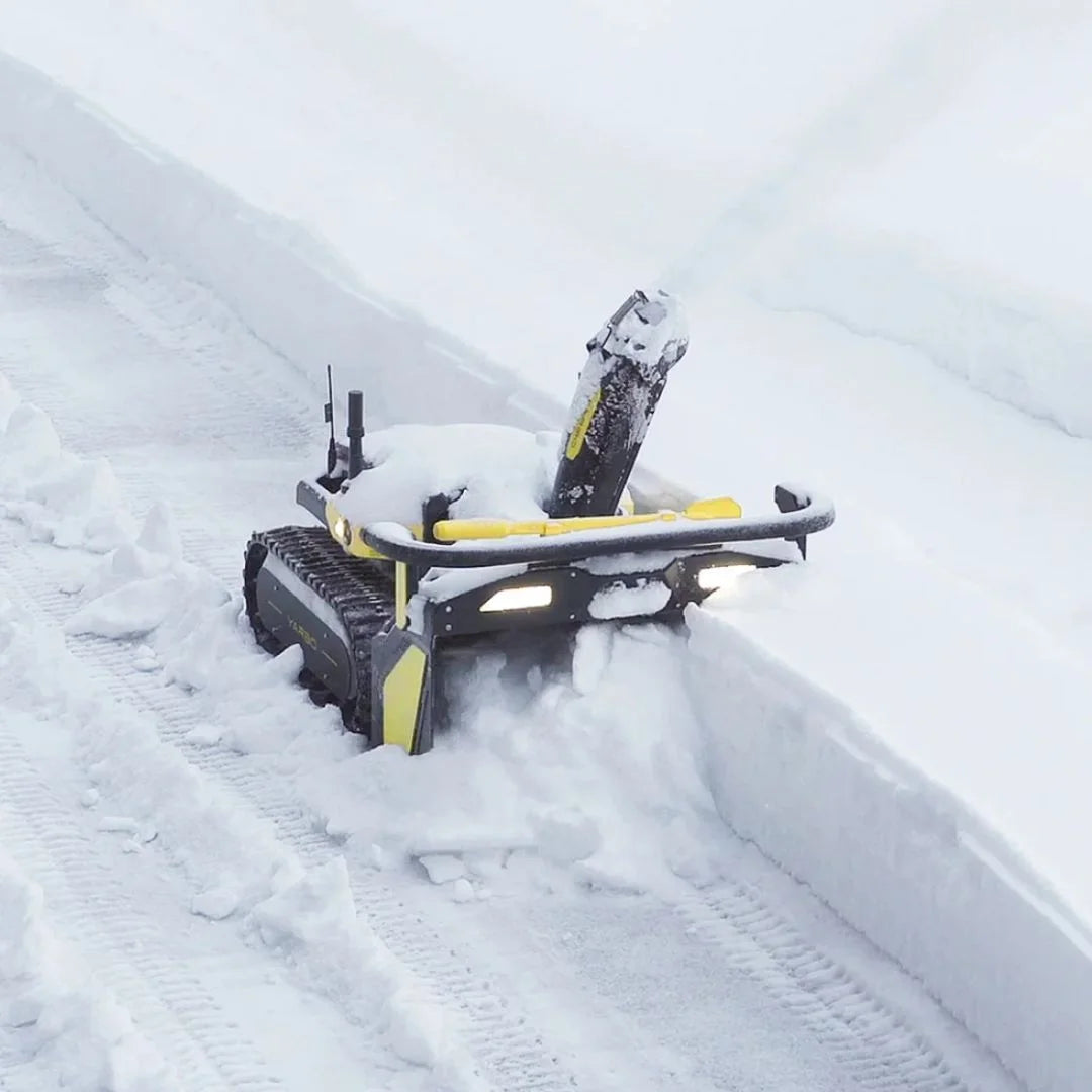 A compact tracked snowmobile or snowcat with yellow accents clearing deep snow in a snowy outdoor setting.