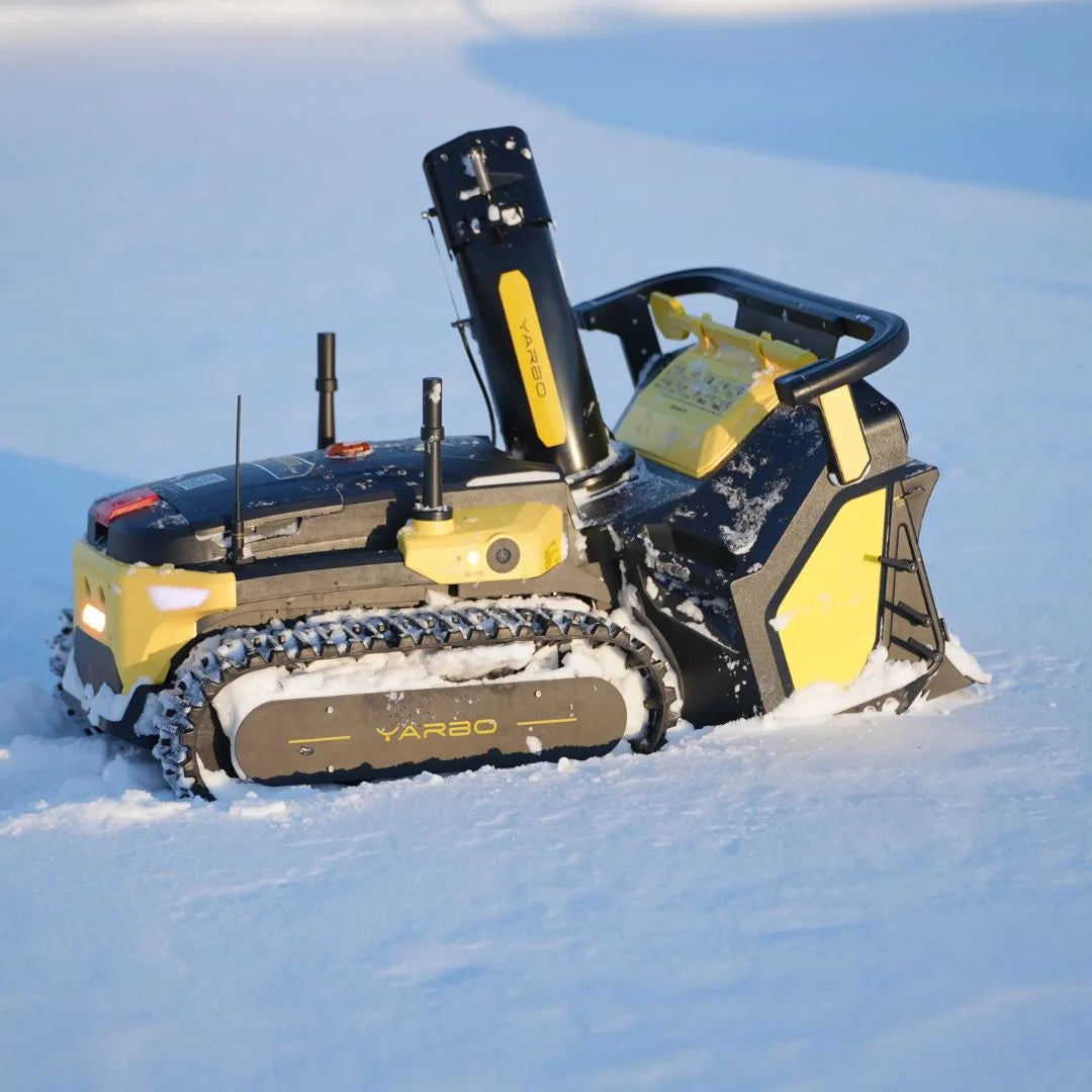 A yellow and black tracked snowmobile or snowcat vehicle driving through deep snow, with visible tracks, a high-mounted operator cabin, and branding that appears to read VARO.